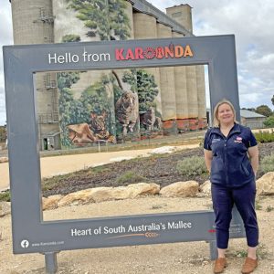 Dee-Anne Hull in front of mural painted silos with a sign saying hello from Karoonda